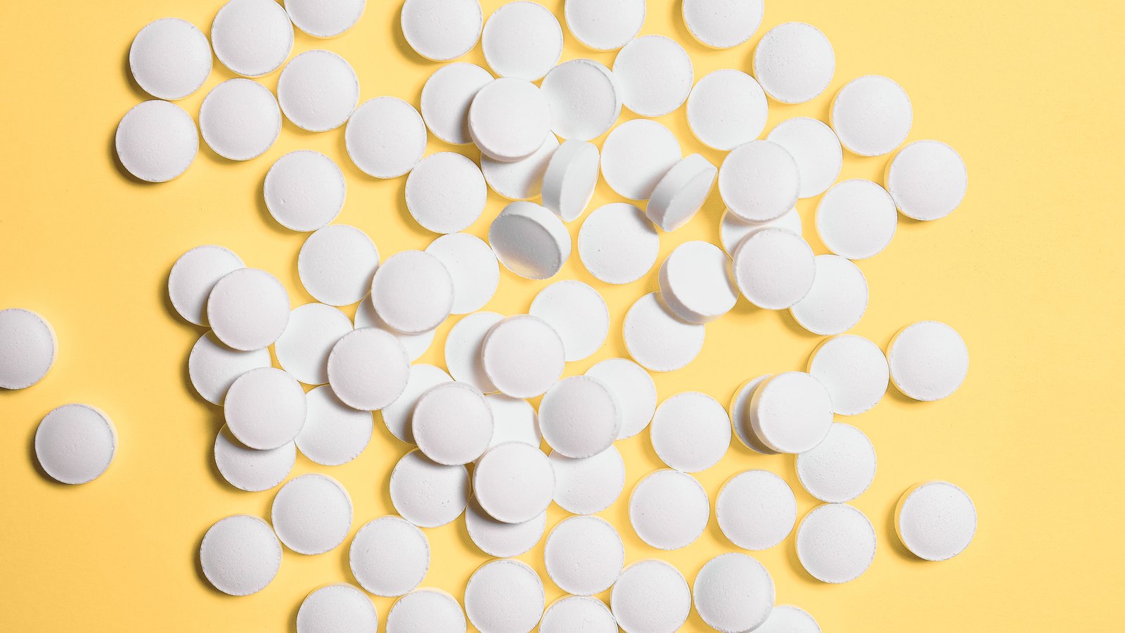 Around a hundred plain white round uncoated tablets scattered loosely across a flat yellow background, unmarked, with no imprint, packaging or lettering anywhere in the frame.