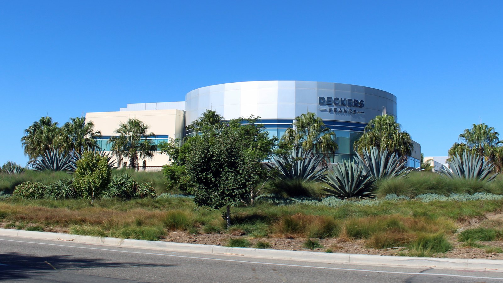 The curved white and glass headquarters building of Deckers Brands in Goleta, California, with its own Deckers Brands lettering, set behind agave plants, palms and a landscaped verge beside a road under a clear blue sky.