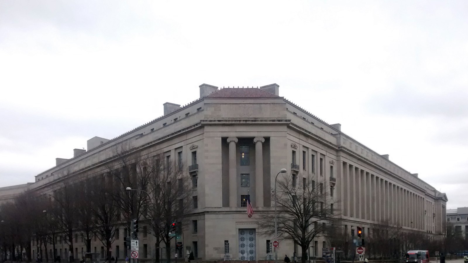 The Robert F. Kennedy Department of Justice Building in Washington photographed from the street corner under a flat overcast sky. A limestone facade runs away to the left and right of the corner bay, with a long colonnade of plain pilasters along the right wing, a tiled roof above the cornice, and an American flag hanging from a pole over the corner entrance. Bare trees and a thin line of street furniture cross the very bottom of the frame.