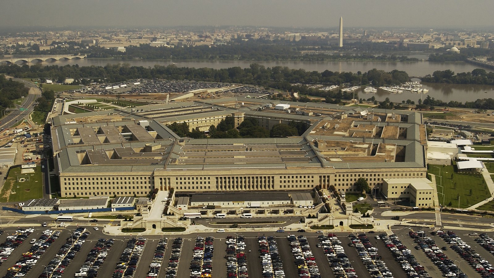 An oblique aerial photograph of the Pentagon, its five sides and inner courtyard visible, with rows of parked cars in the foreground and the Potomac, the Washington Monument and the city beyond.