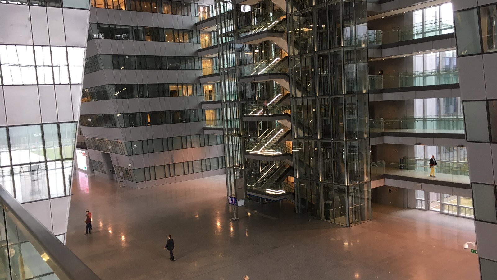 The central atrium of the NATO headquarters building in Brussels, with a glazed tower of escalators rising through it and a few people crossing the stone floor below.
