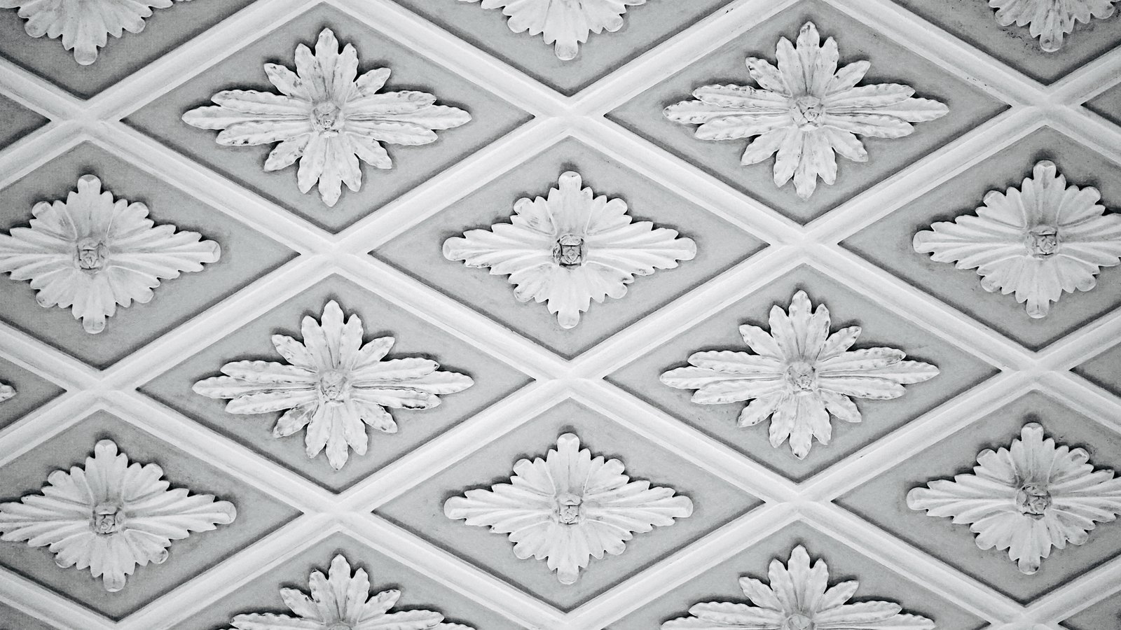 A close photograph of a moulded grey and white ceiling panel, raised ribs dividing it into a diamond lattice with a carved rosette centred in every diamond. No text, brand mark, person or place is in view.