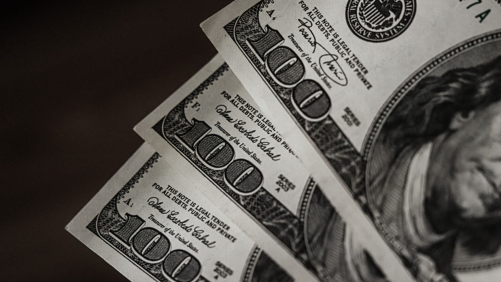 Three United States one hundred dollar bills fanned across each other in black and white, the 100 numerals, the legal tender line and part of the Franklin portrait in frame against a dark background.