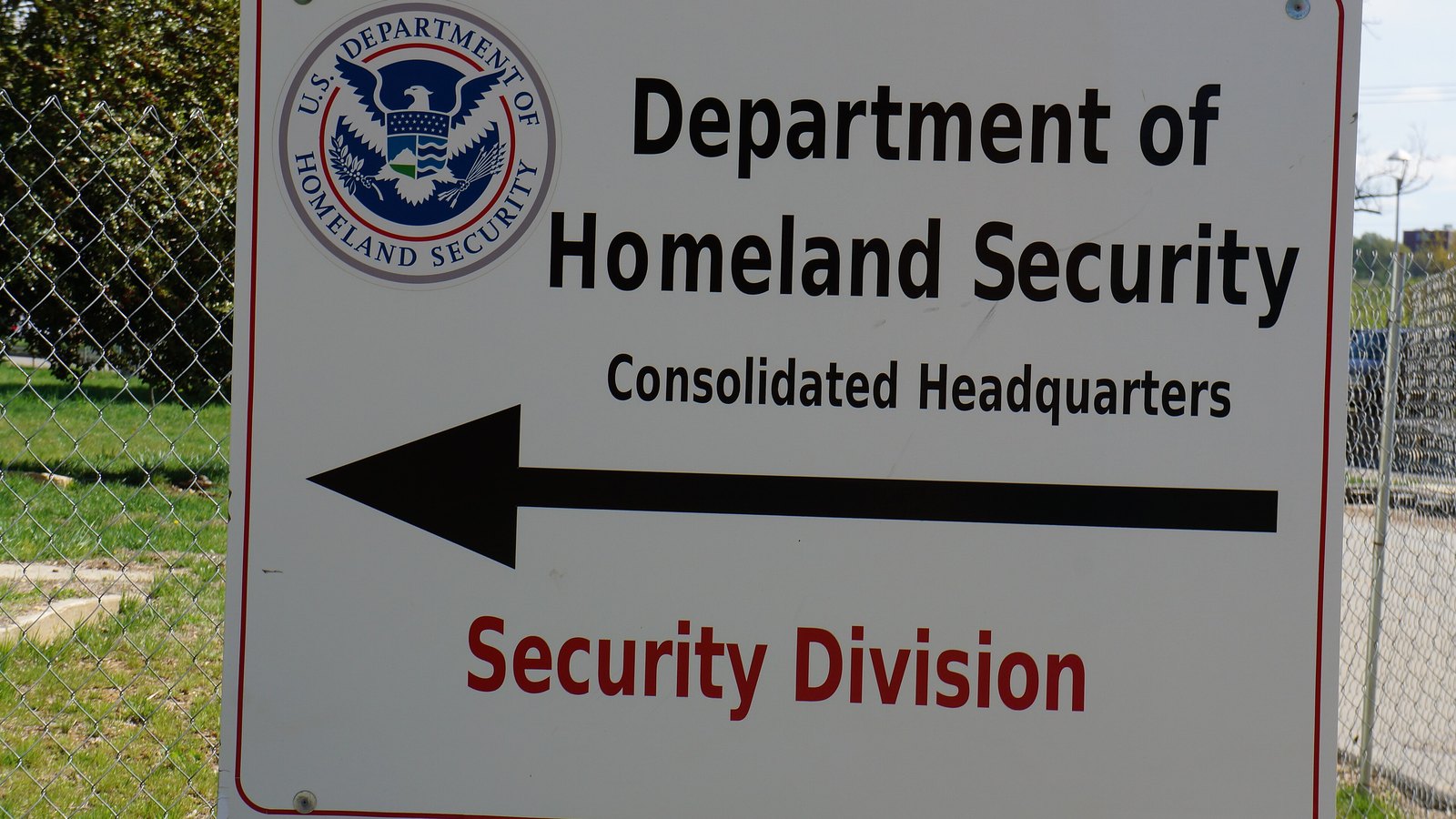 A white sign with the Department of Homeland Security seal reading Department of Homeland Security Consolidated Headquarters, with an arrow pointing left and the words Security Division, mounted in front of a chain-link fence and grass.