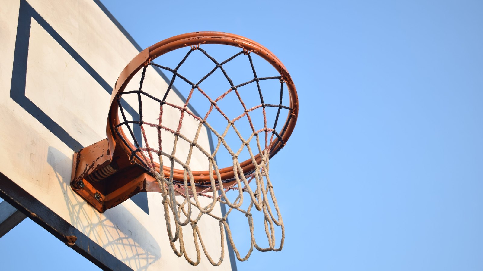 An outdoor basketball hoop photographed from below, the orange rim chipped and worn, the net a mix of dark and pale cord, mounted on a plain white backboard against a clear blue sky.