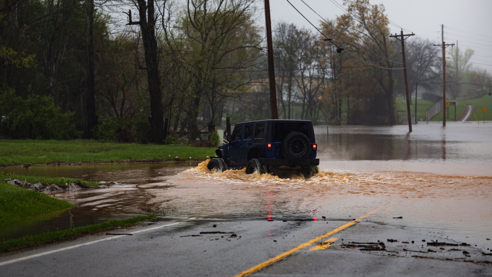 A dark Jeep drives through floodwater covering a tree-lined road, not a photograph of this event.