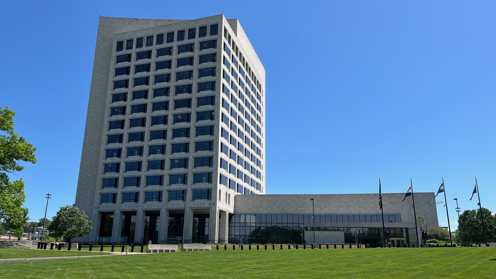 The Federal Reserve Bank of Kansas City, a pale limestone tower above a low colonnaded entrance wing, seen from across the street under a clear blue sky, with the bank's name cut into the stone frieze and United States and state flags flying on the forecourt.