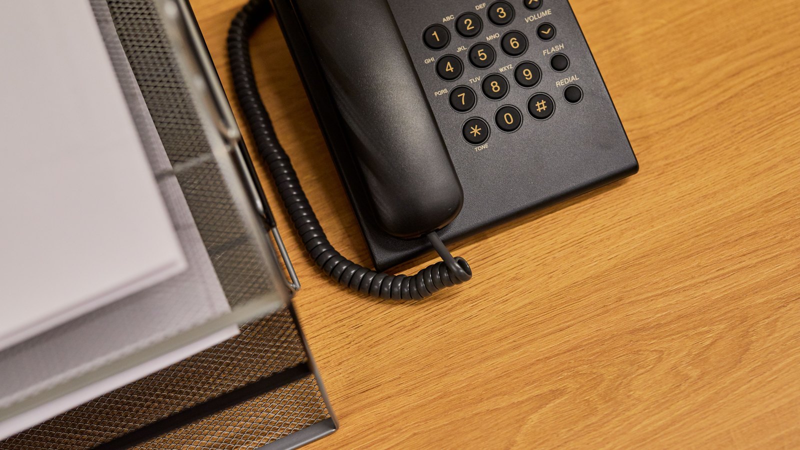 A photograph illustrating a plain black corded desk telephone with the handset lying off the hook on a bare desk.