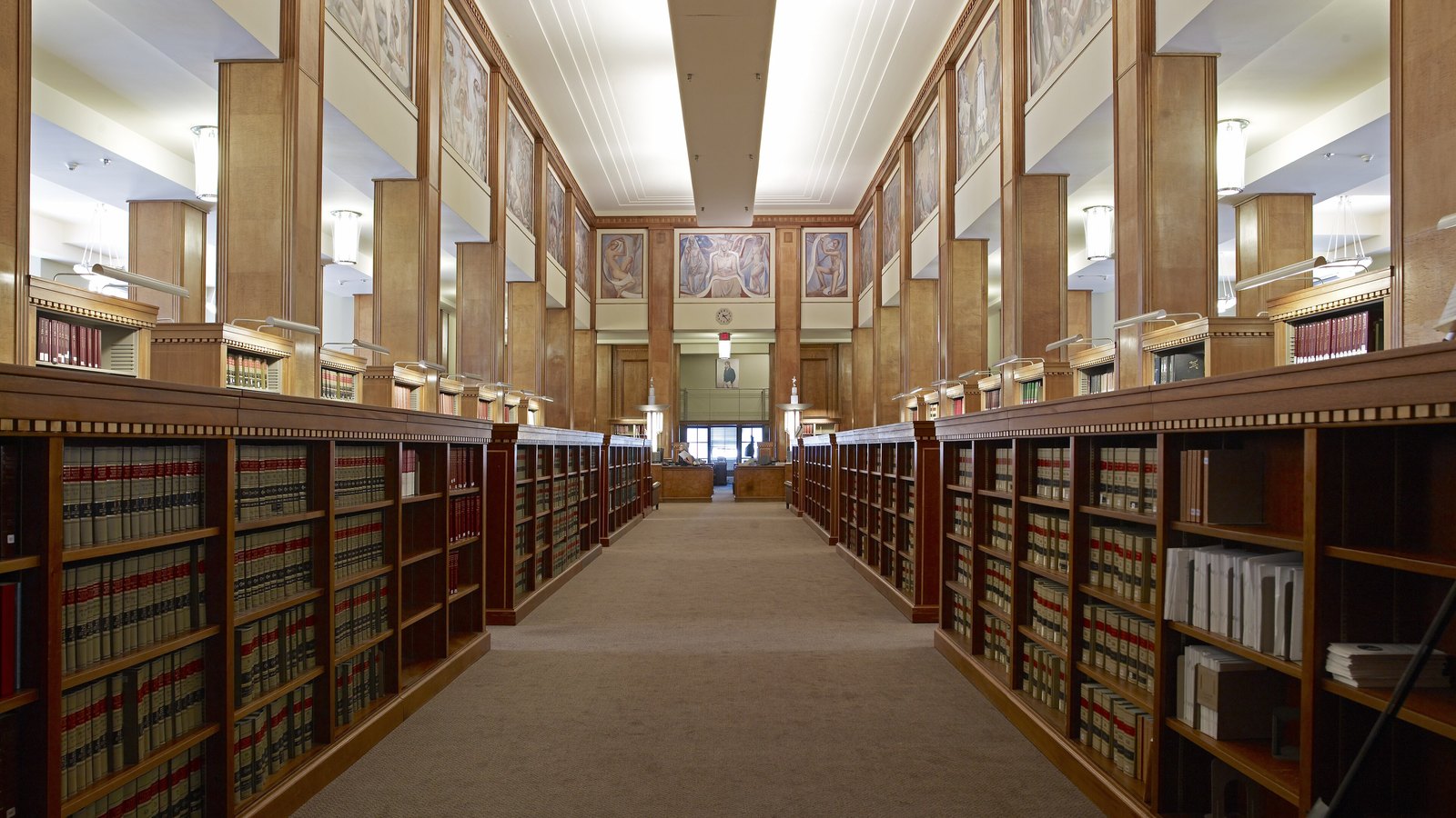 The main library inside the Robert F. Kennedy Department of Justice Building in Washington, D.C., looking down a carpeted aisle between long runs of shelved bound law reports towards a mural at the far end.