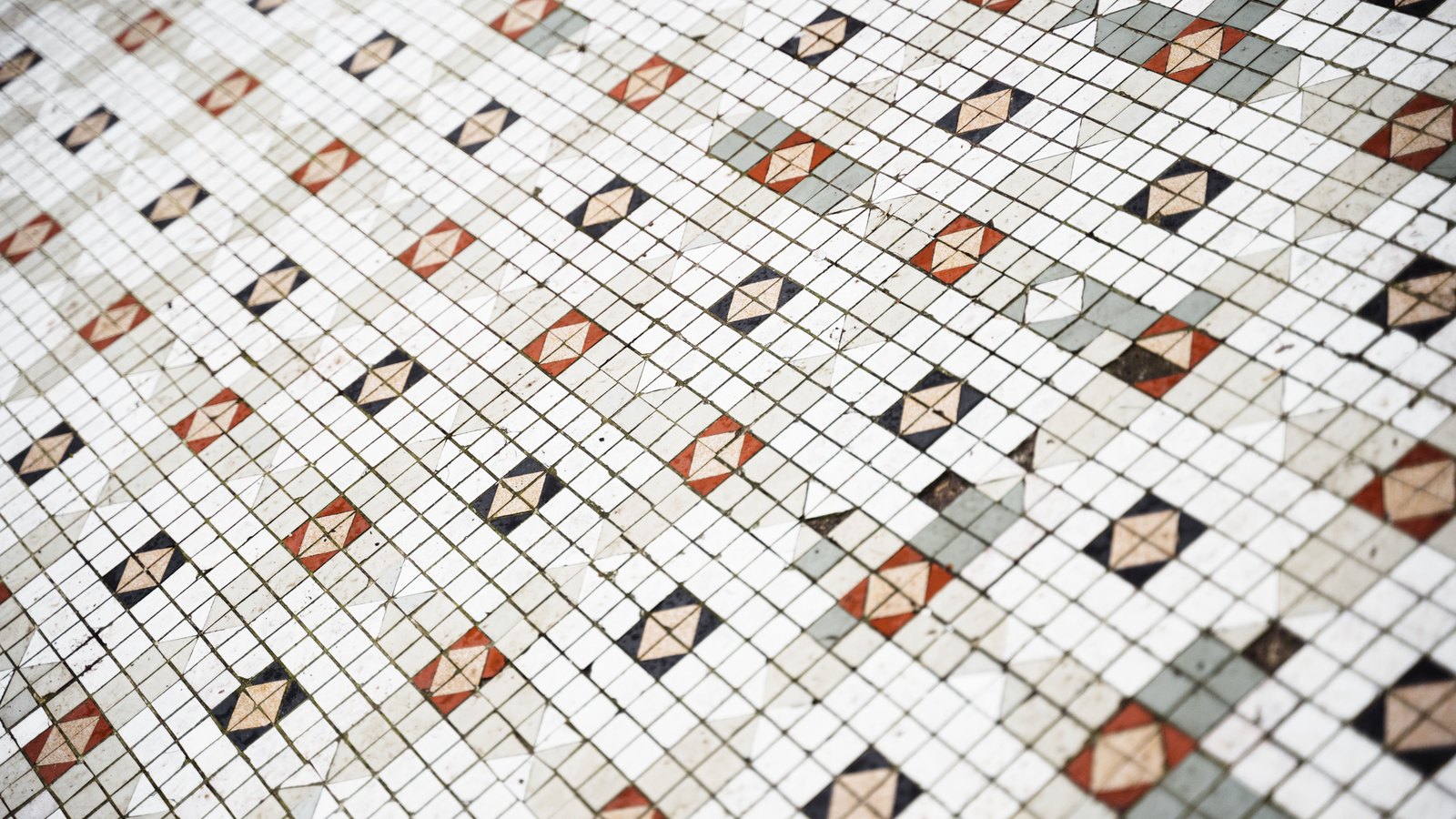 An old mosaic floor seen at an angle, laid in small square white tiles with a scattered repeating motif of red, navy and pale sage diamonds set into the grid. The joints are dark with age. No horizon, wall, doorway, furniture, person or lettering is in the picture.