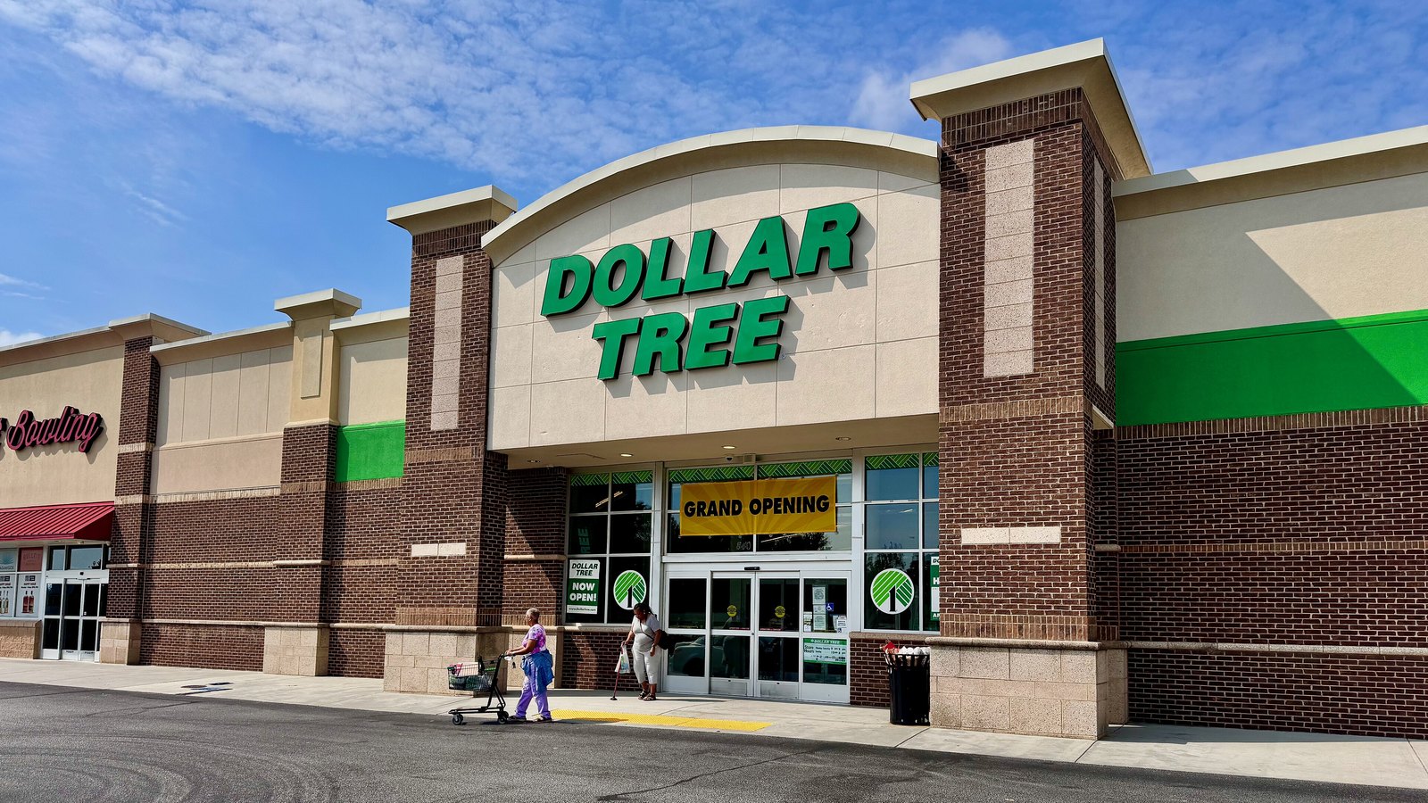 The front of a Dollar Tree store in Greenville, South Carolina, seen from the car park under a blue sky: a low brick and stucco building with the DOLLAR TREE wordmark in green capitals on the raised centre section, glass entrance doors beneath it carrying a yellow GRAND OPENING banner, two shoppers walking out with a trolley, and the edge of a neighbouring bowling unit at the left.