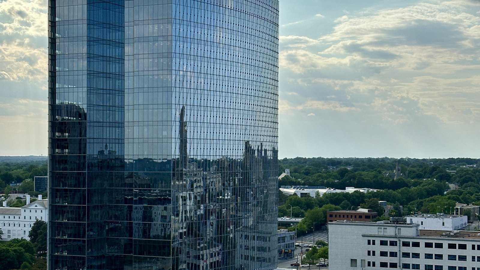 The glass curtain wall of 600 Canal Place, the Dominion Energy headquarters tower in Richmond, Virginia, photographed from a neighbouring rooftop, with the city and its treeline reflected across the face of the building and low office blocks and a highway laid out below.