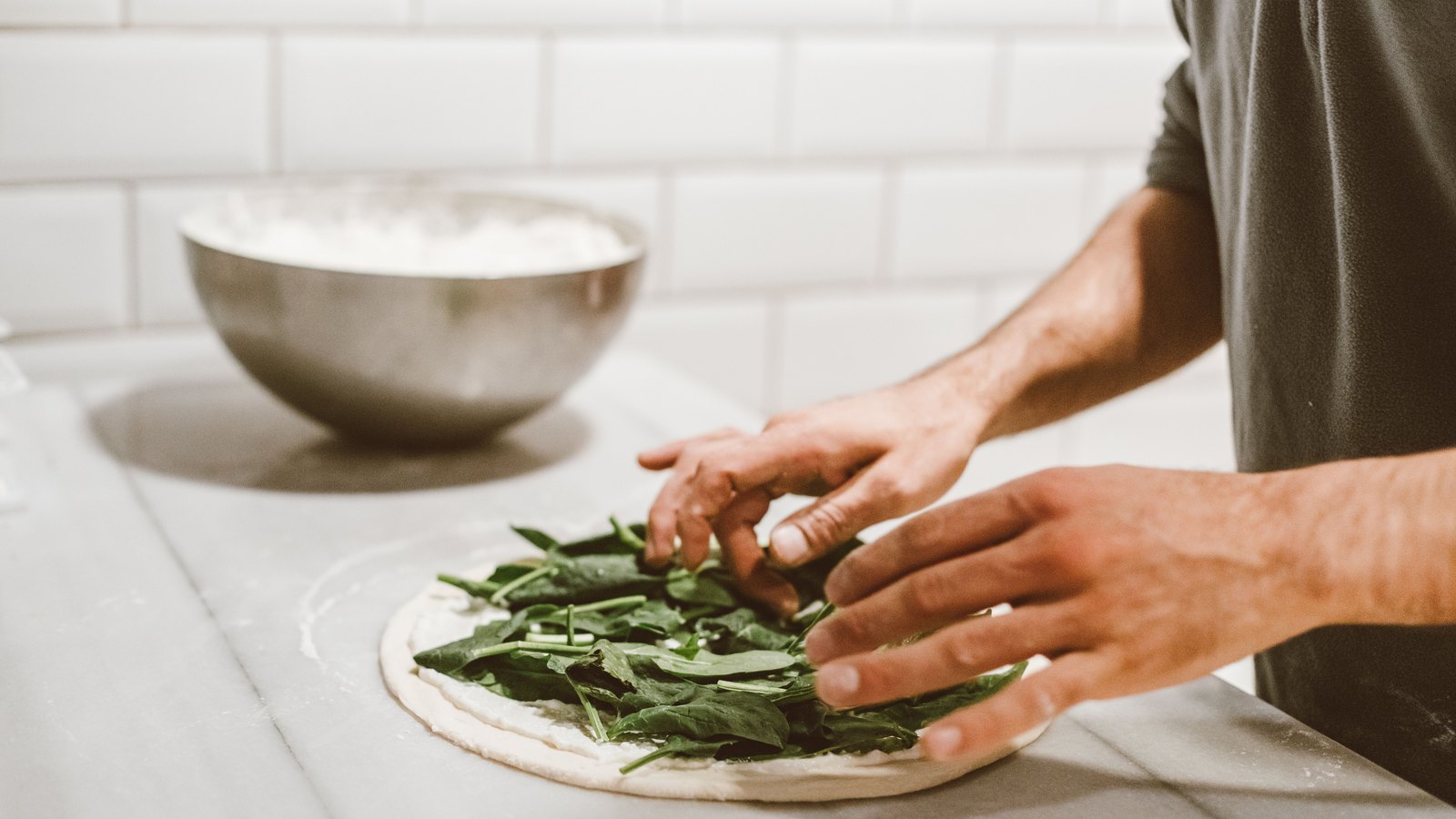 A person's hands arranging spinach leaves on a round of raw pizza dough, with a metal bowl of flour on a marble counter behind.