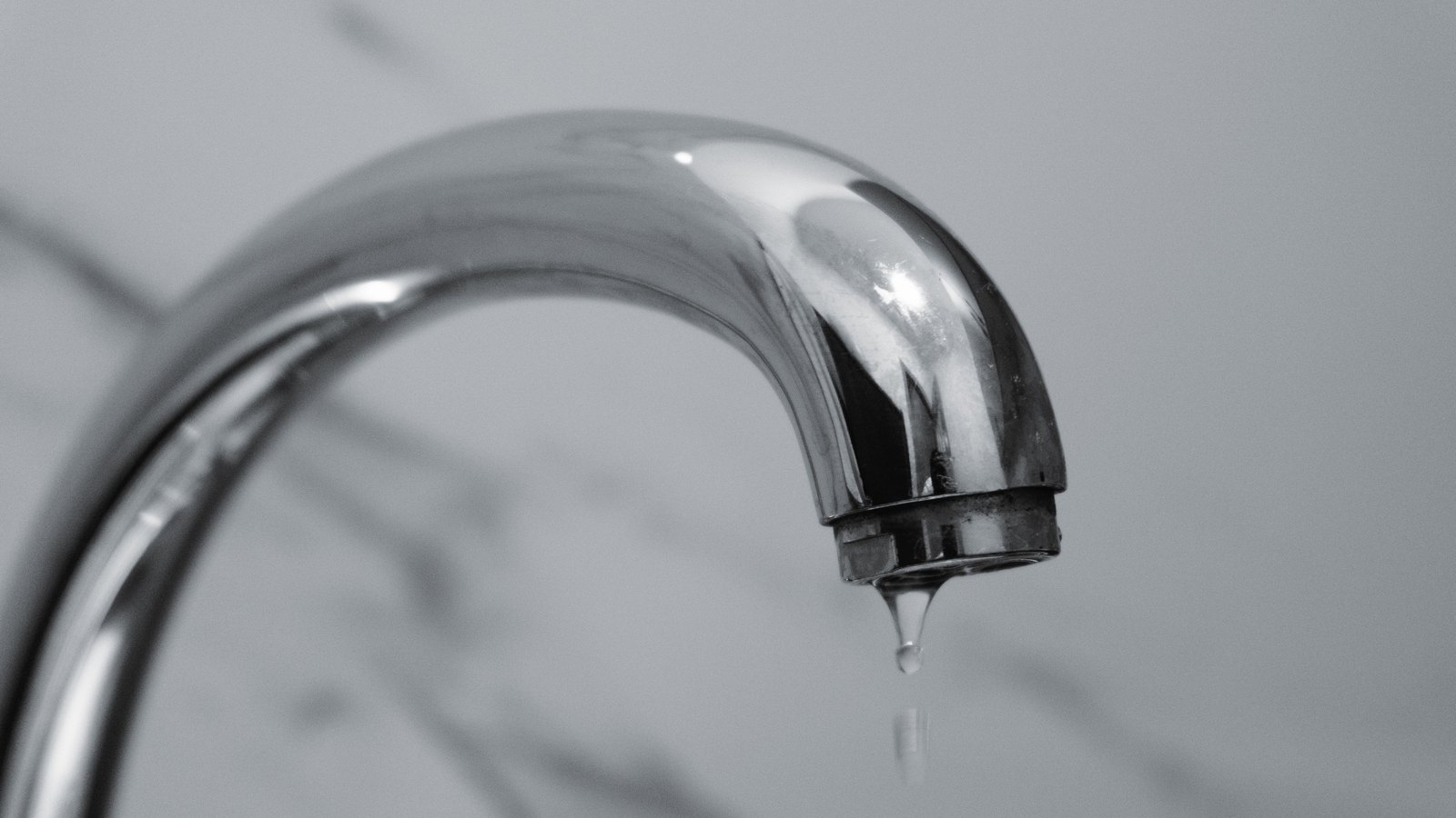 A close-up black and white photograph of a curved chrome tap spout against a plain pale background, with a single drop of water hanging from the aerator at its tip. No people, hands, rooms, addresses, signs, branding or text appear anywhere in the frame. Stock photograph, not a photograph of this event.