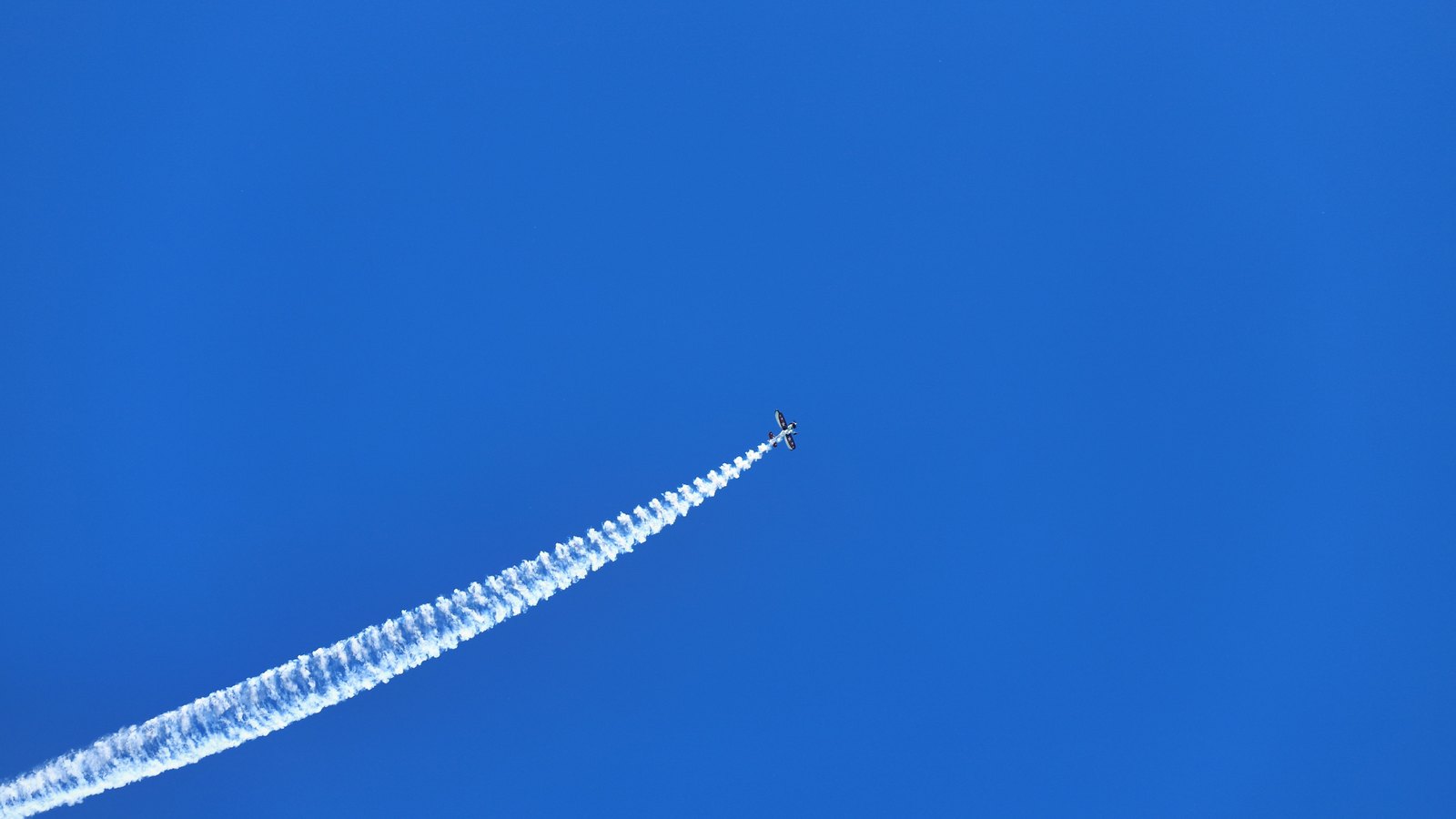 A single aircraft, small and dark at high altitude, crossing a deep blue cloudless sky and trailing a long white vapour trail that runs diagonally out of the bottom left of the frame.