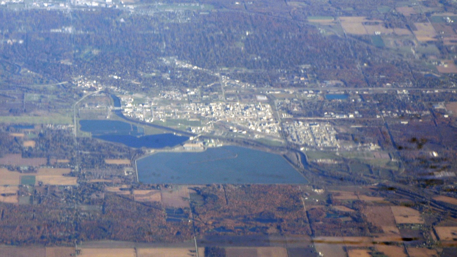 An aerial view of Midland, Michigan, showing the pale buildings of the Dow Chemical plant and headquarters beside large blue brine ponds, surrounded by autumn woodland and farm fields.