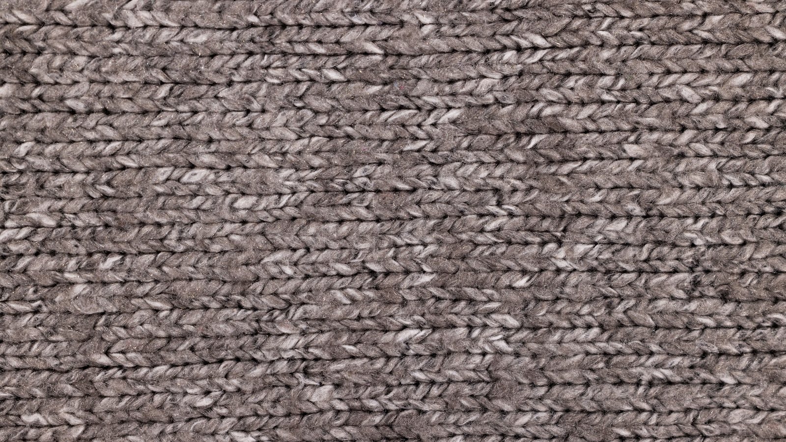 A close photograph of coarse knitted wool filling the whole frame, ranks of chunky taupe and grey stitches running left to right in even rows, the yarn flecked lighter and darker where the fibres twist. No text, brand mark, person or place is in view.