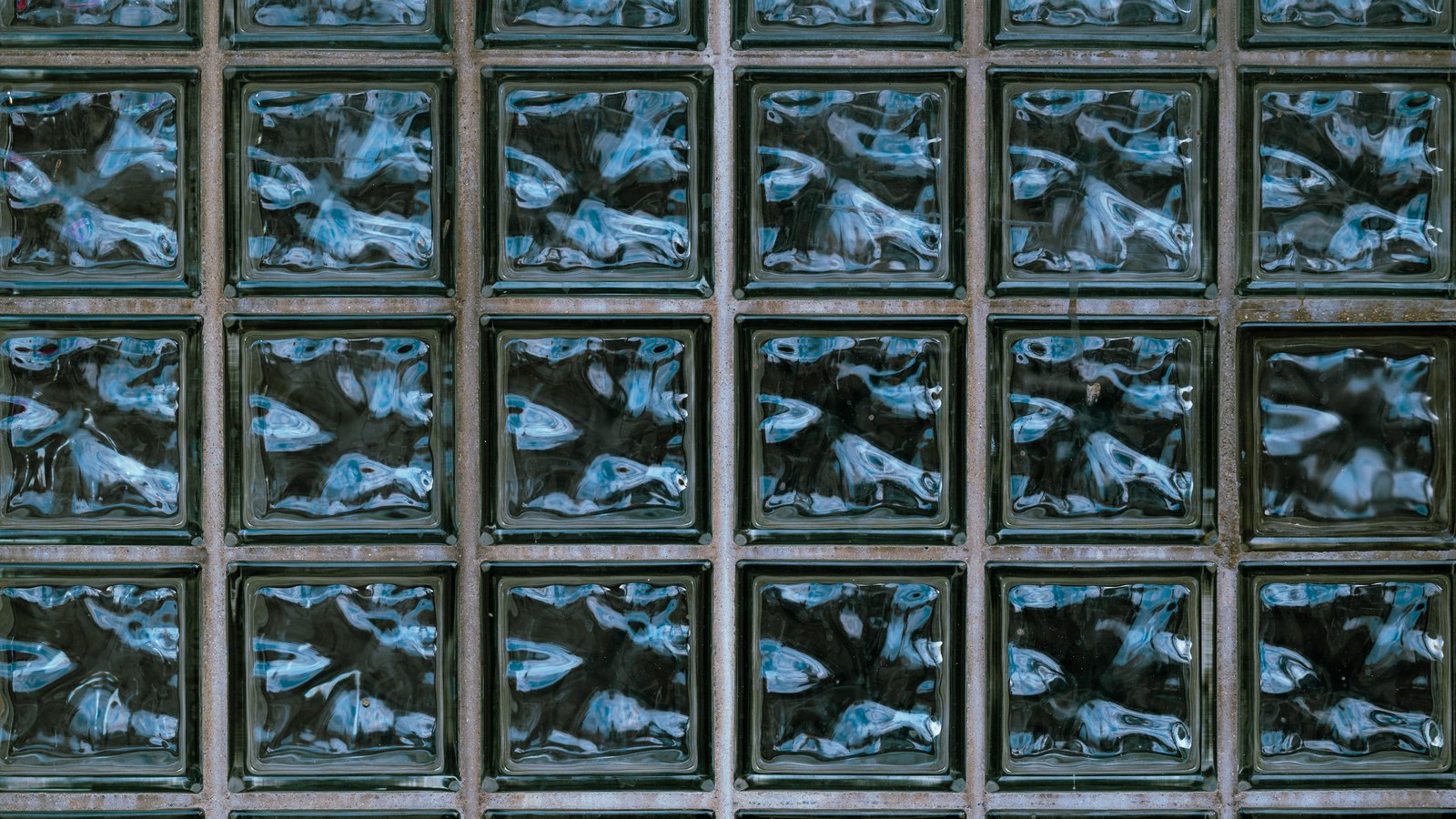 A close photograph of a wall of square glass blocks laid in four courses and filling the frame. Each block is moulded with a rippled interior that scatters light into pale blue and white streaks against a near black ground, and narrow weathered mortar joints run between them. No text, markings, brand marks, people or machinery are in view. A generic architectural glass close-up, not a photograph of any power station, reactor or plant site.