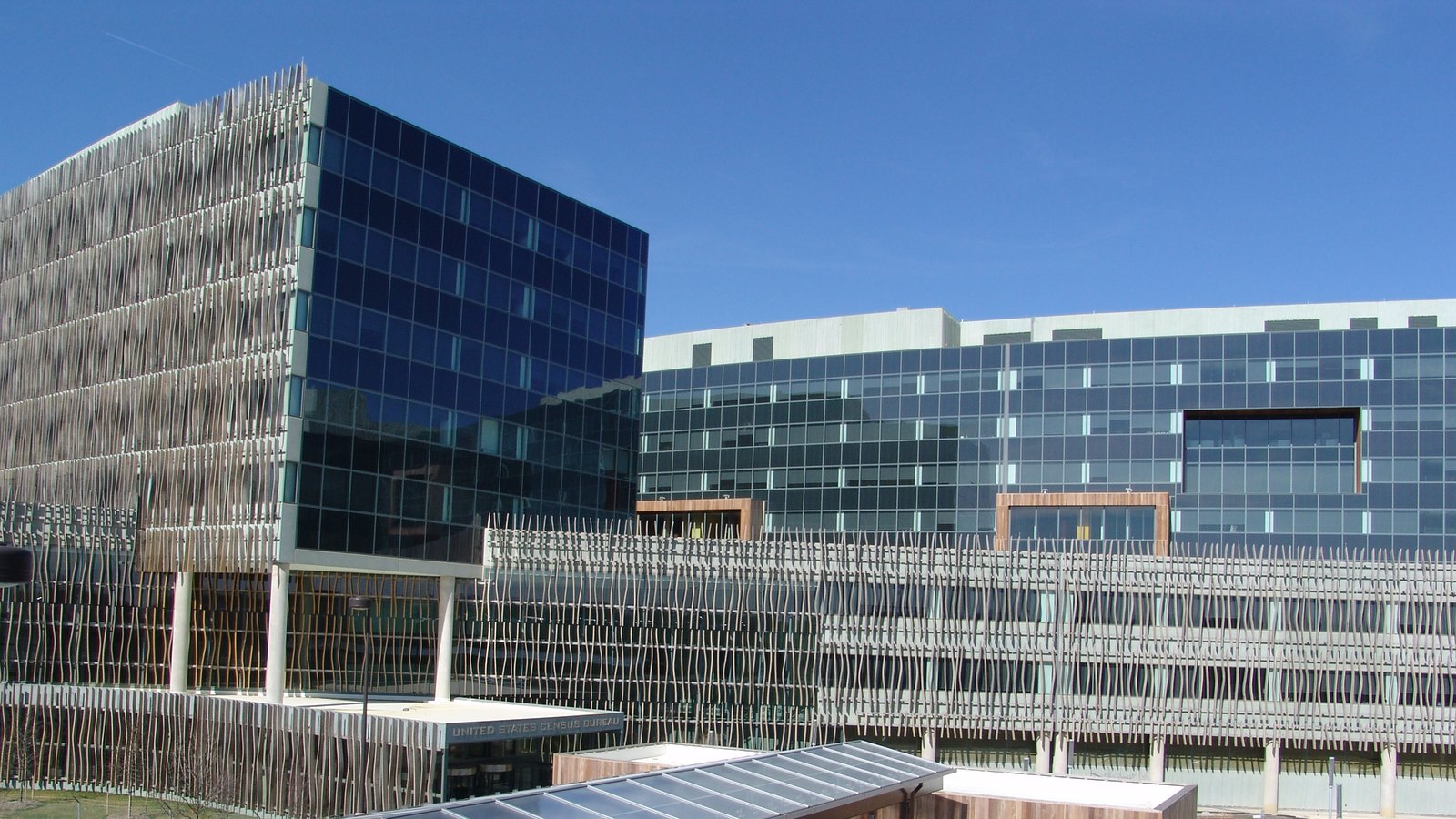 The United States Census Bureau headquarters at Suitland, Maryland: a dark blue glazed corner block joined to a longer pale grey wing, both screened along the front by a curved lattice of vertical metal blades, with the words UNITED STATES CENSUS BUREAU on the entrance canopy at the foot of the frame, under a clear sky.