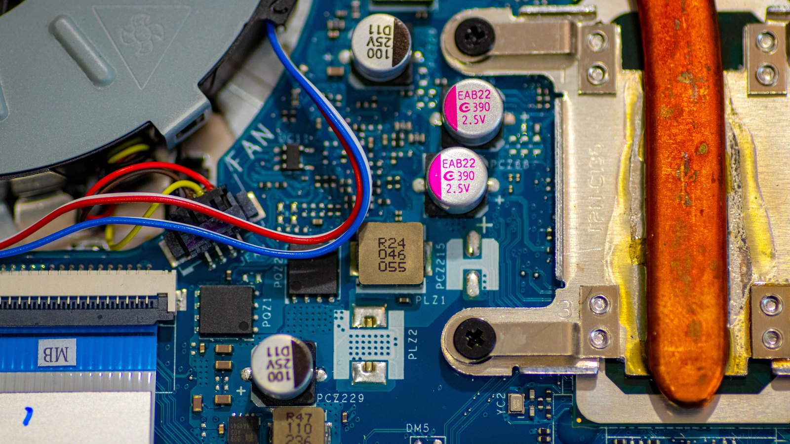 Close view inside a laptop, showing a blue circuit board dense with capacitors and surface mount components, a copper heat pipe running down the right side, and red, blue and yellow fan wires crossing the upper left.