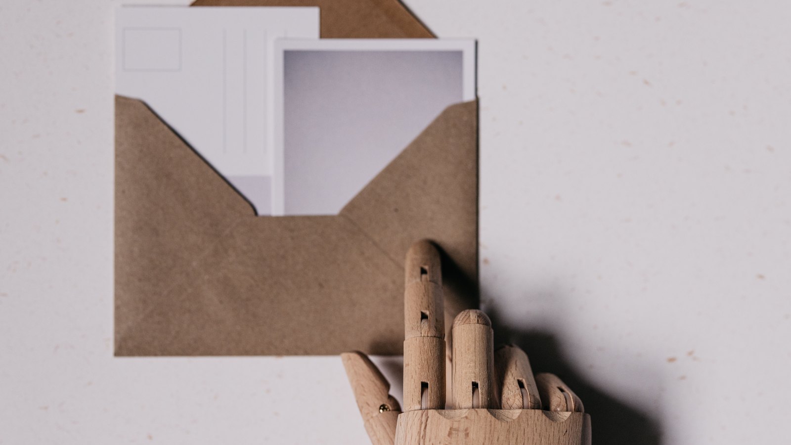 A studio photograph looking down on an open kraft paper envelope lying on a pale speckled surface, with two blank white cards part way out of it. A jointed wooden artist's mannequin hand rises from the lower edge of the frame with one finger extended toward the envelope, casting a soft shadow to the right. No people, no lettering and no place can be identified from it.