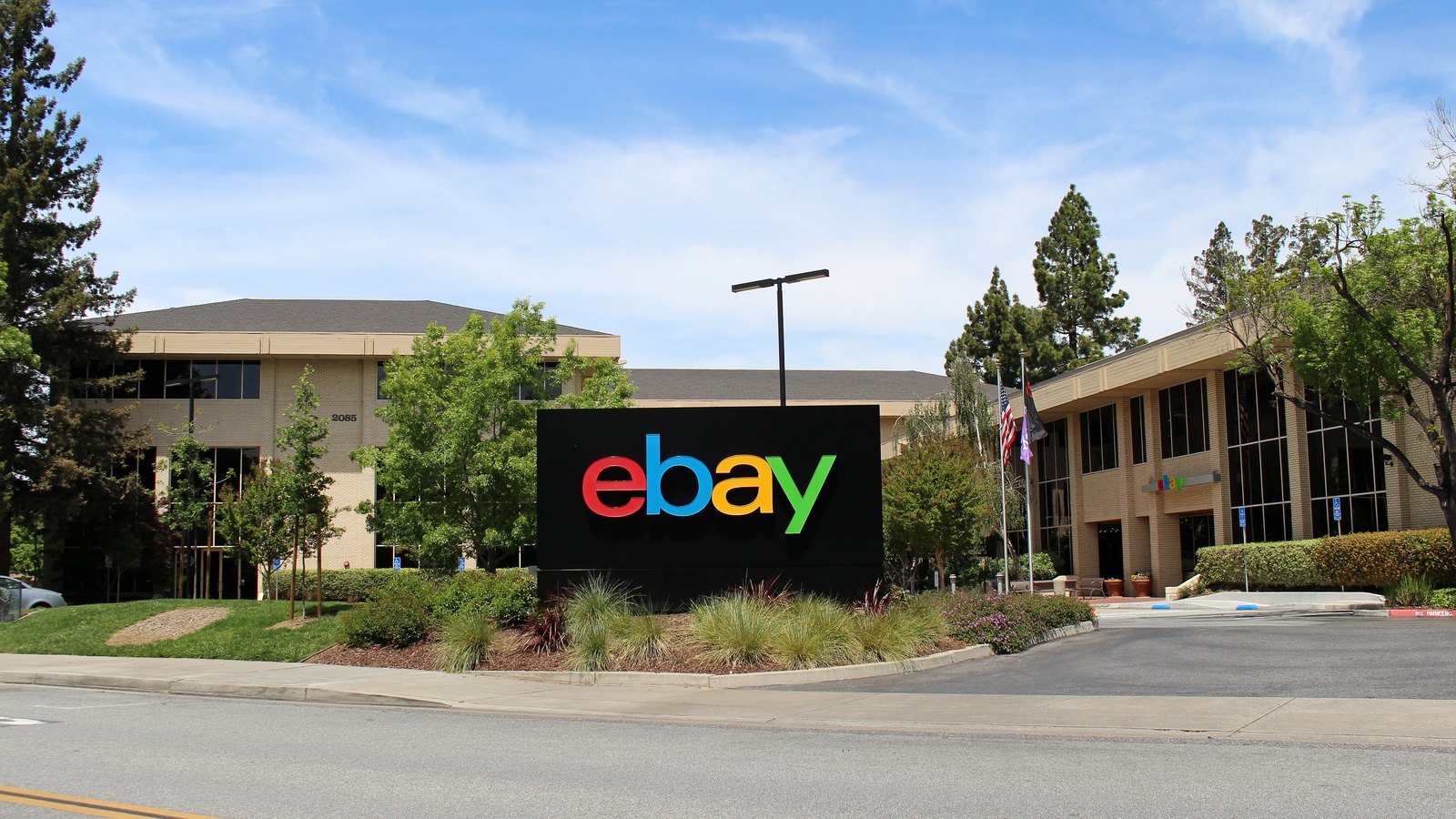 A black monument sign bearing the multicoloured eBay logo in front of low beige office buildings at the company's San Jose headquarters, under a blue sky with tall pines.