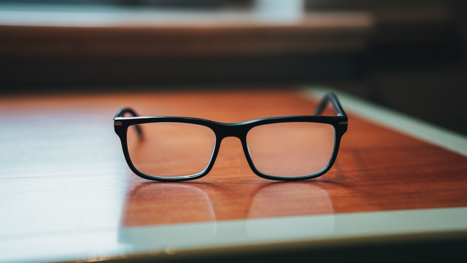 A pair of black framed spectacles standing open on a dark wooden table beside the pale edge of a closed folder, with a warm, blurred interior behind them. No text, brand mark or person is in the frame.