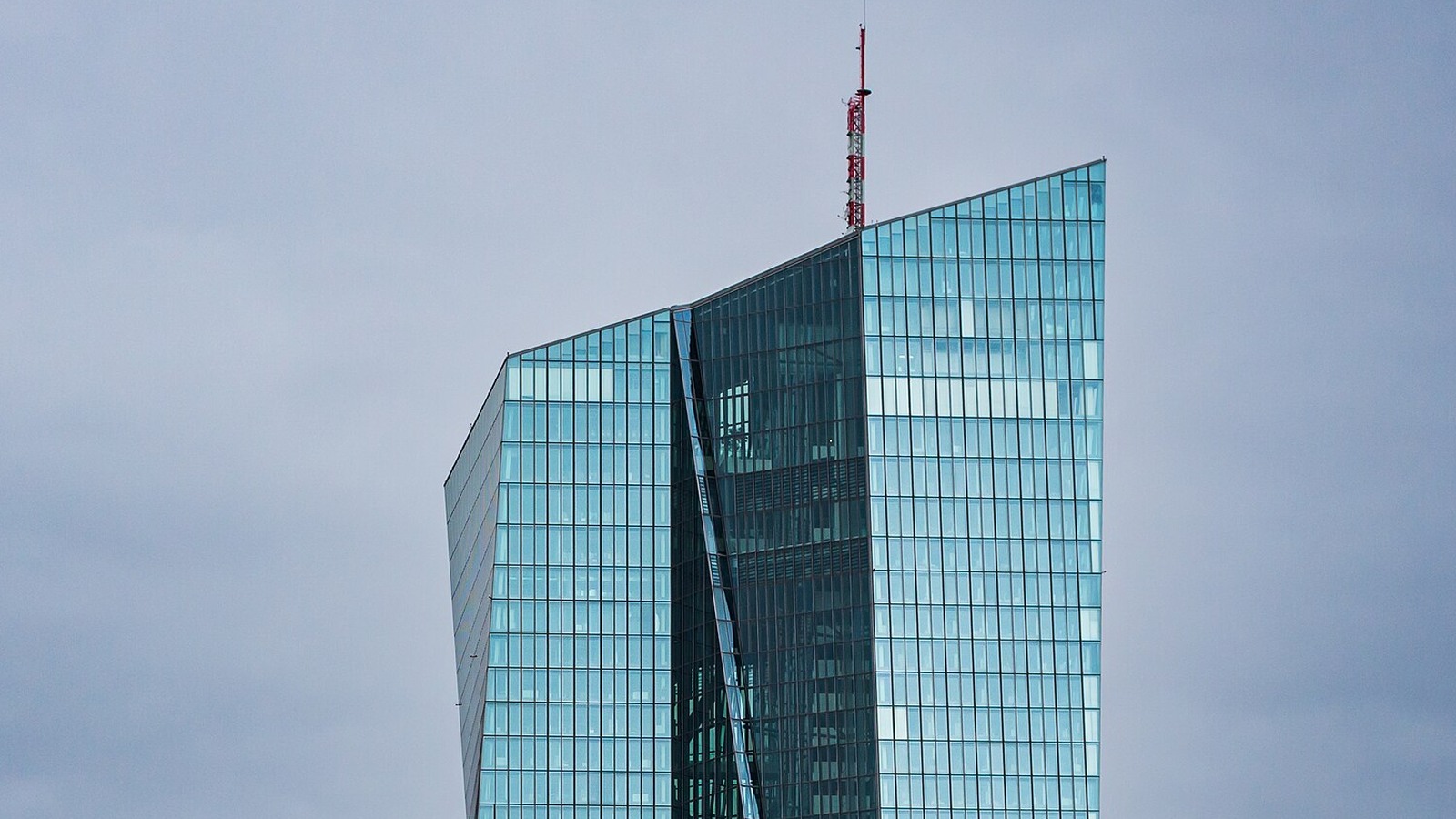 The upper glass facades of the European Central Bank headquarters in Frankfurt against a cloudy sky.