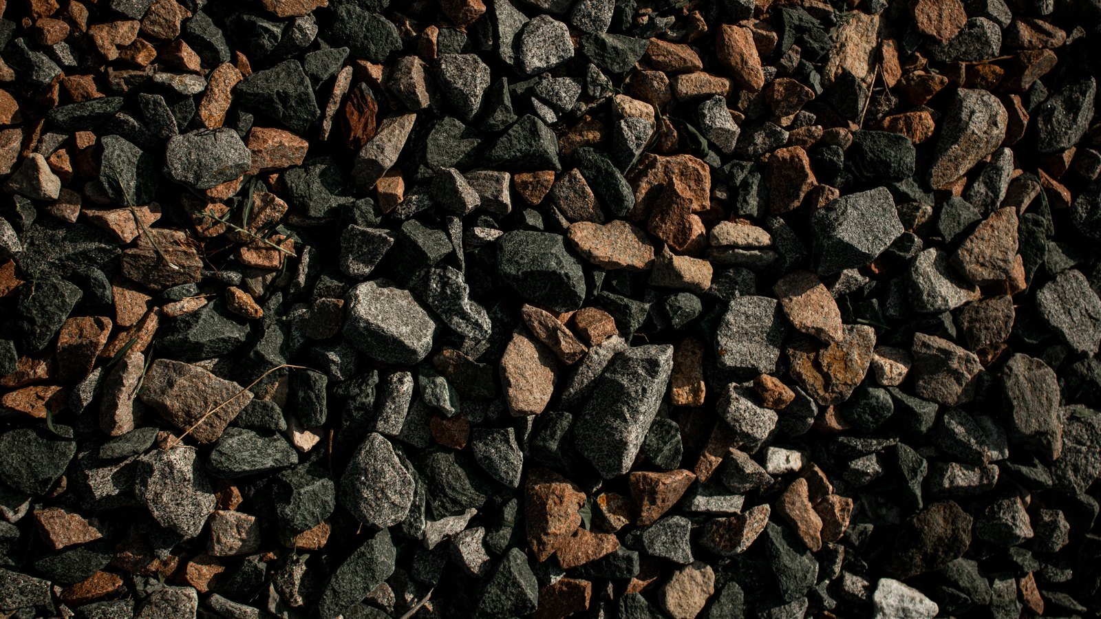 A close overhead crop of coarse crushed stone, angular grey and rust brown chippings packed tight across the whole frame in low raking light, with a few dry grass blades caught among them. There is no road marking, no lettering, no signage, no vehicle and no person in the frame, and no place can be identified from it. A generic illustration of road aggregate, not a photograph of the Edison Bridge, of the repair plate or of any damage to anyone's vehicle.