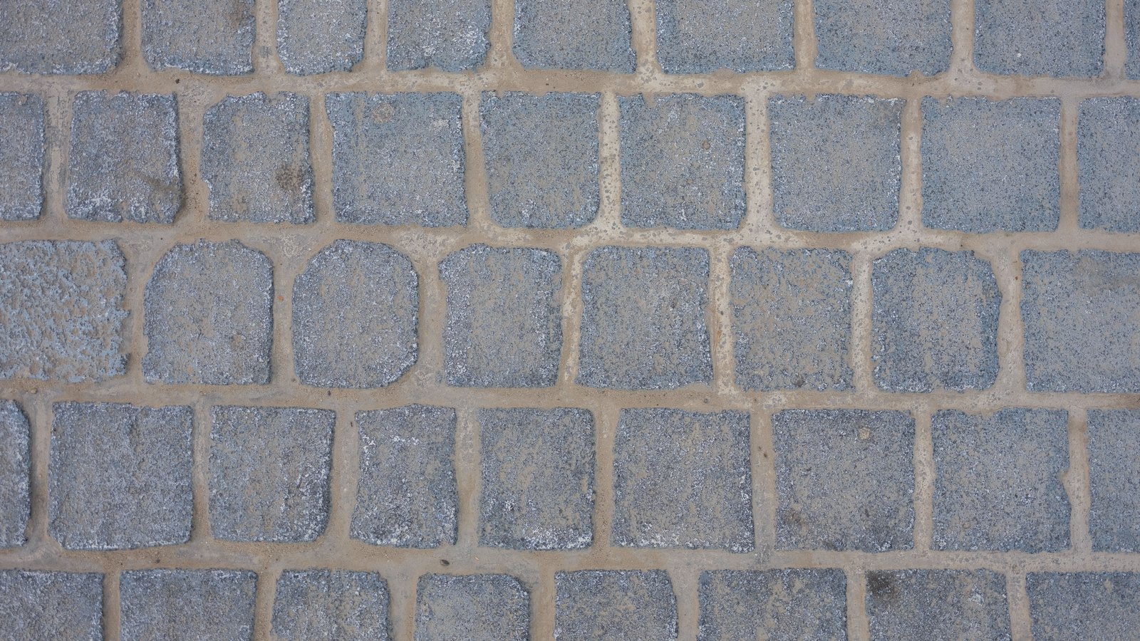 A square-on overhead crop of a paved surface of grey stone setts laid in irregular courses, each block coarse and speckled and separated from its neighbours by wide pale sandy joints. The pattern fills the frame. No text, brand mark, person, kerb or horizon is in view.