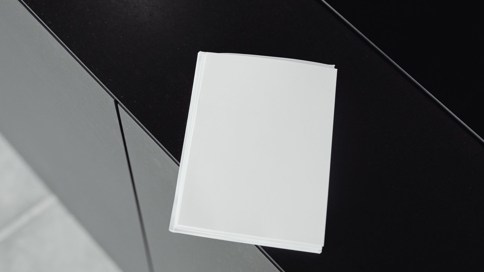 An overhead view of a squared-off stack of blank white paper sheets resting on a dark matt surface, lit so the edges of the individual pages separate, with a plain grey floor at the edge of the frame. No person, logo, letterhead, agency name or legible text of any kind appears anywhere in the shot. A generic illustration of a long printed notice, not a photograph of any actual document.