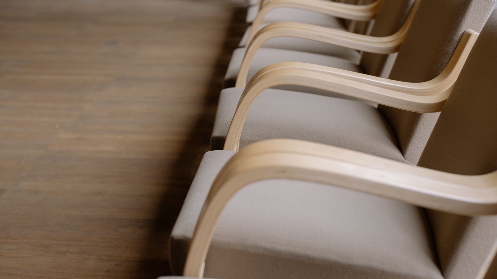 A close crop of a row of empty armchairs with pale bentwood arms and grey upholstery, receding down a bare wooden floor in soft daylight.
