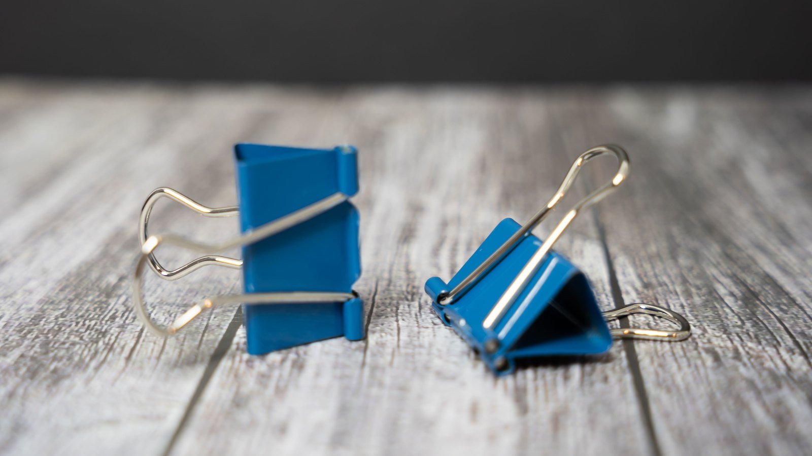 Two blue steel binder clips standing on a pale whitewashed wooden surface against a dark grey background, one seen edge on and one from the front.