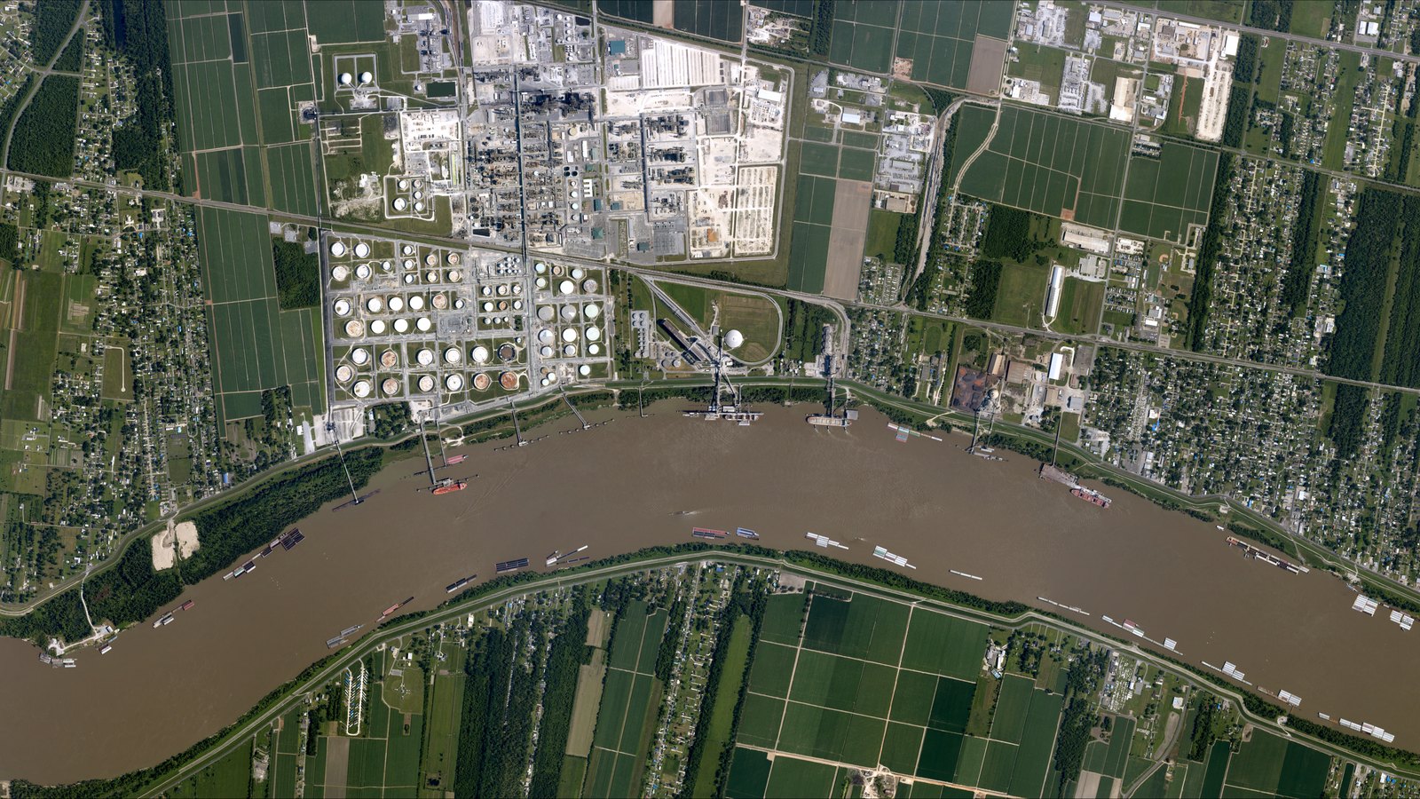 A vertical aerial photograph of a large oil refinery on a bend of the Mississippi River: process units and industrial buildings across the upper half, a grid of roughly sixty white and rust coloured storage tanks below them, a white sphere and loading docks at the water's edge with barges moored along the brown river, and cane fields and residential streets running back from both banks.