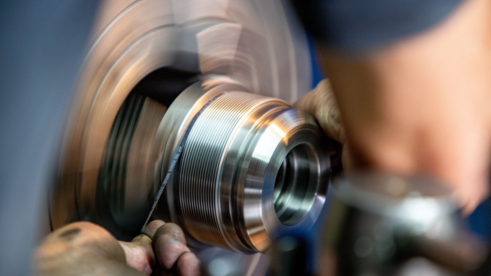 A metal component turning on a lathe, its bright machined threads and bore held in sharp focus while the spinning disc behind blurs into motion, a worker's hands steadying tooling at either side, with no lettering or branding in view.
