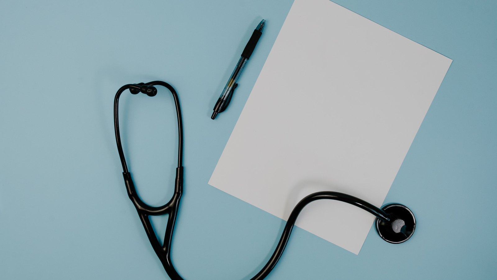 A black stethoscope and a pen beside a blank white sheet of paper on a pale blue surface.