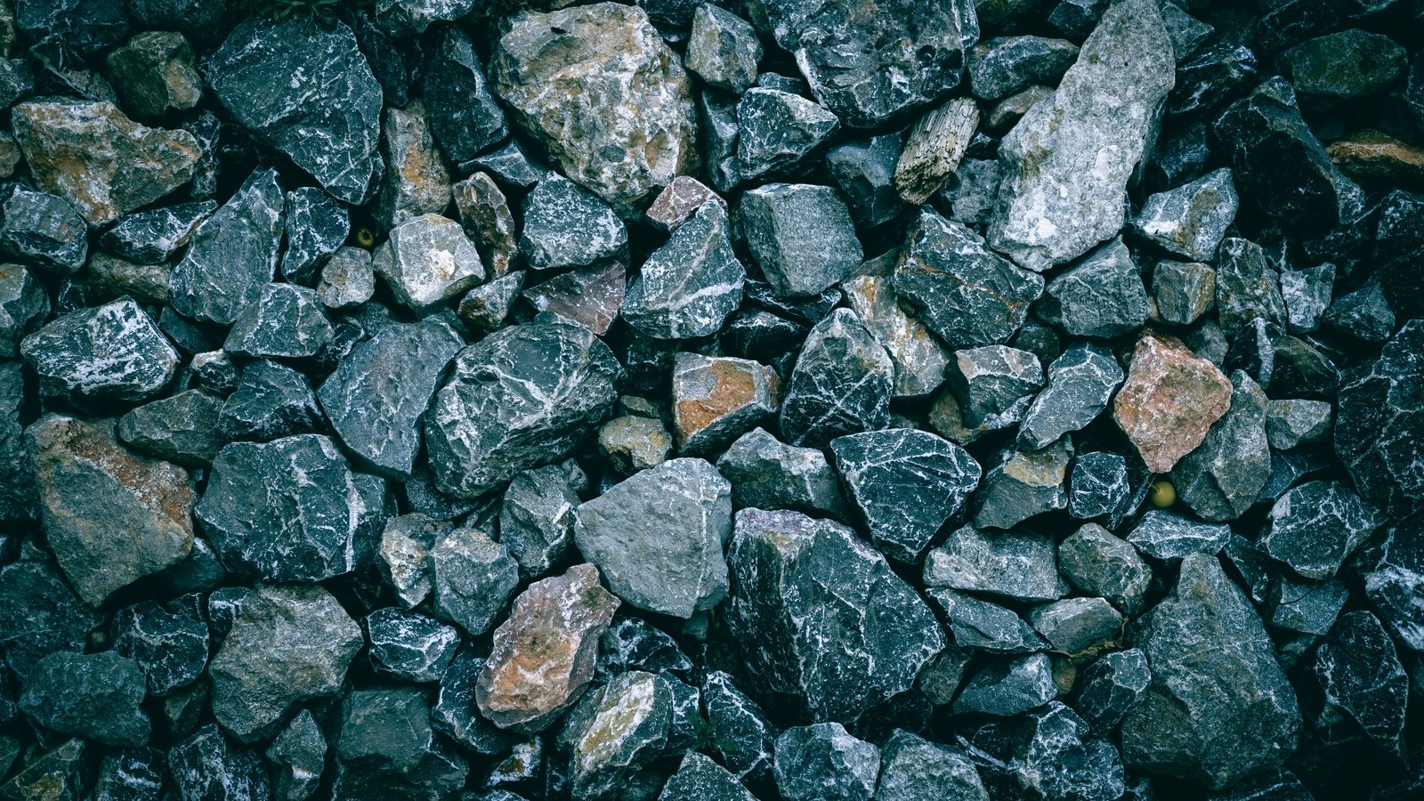 A top-down close photograph of a bed of angular crushed stone filling the frame, most pieces dark blue-grey with white quartz veining and a few ochre and pink fragments, with no tool, hand or marking in the picture.