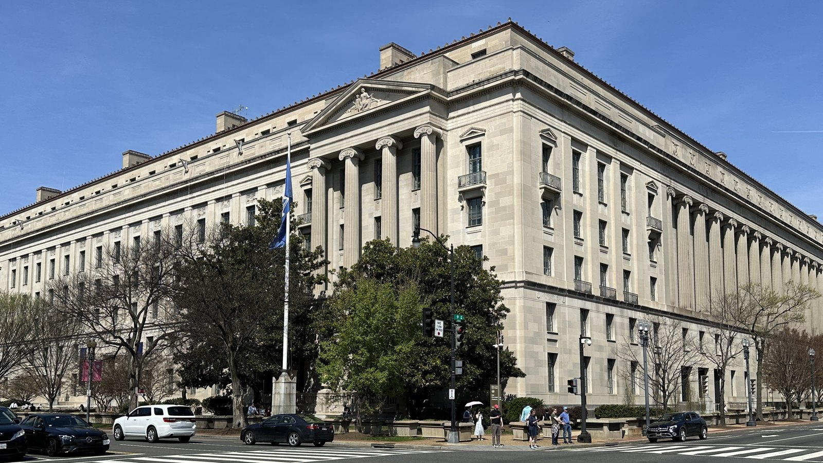 The Robert F. Kennedy Department of Justice Building in Washington, photographed from the far side of the street under a clear blue sky. Two limestone elevations of fluted columns meet at a corner beneath a sculpted pediment, with a blue flag on a pole, street trees and passing traffic in the foreground.
