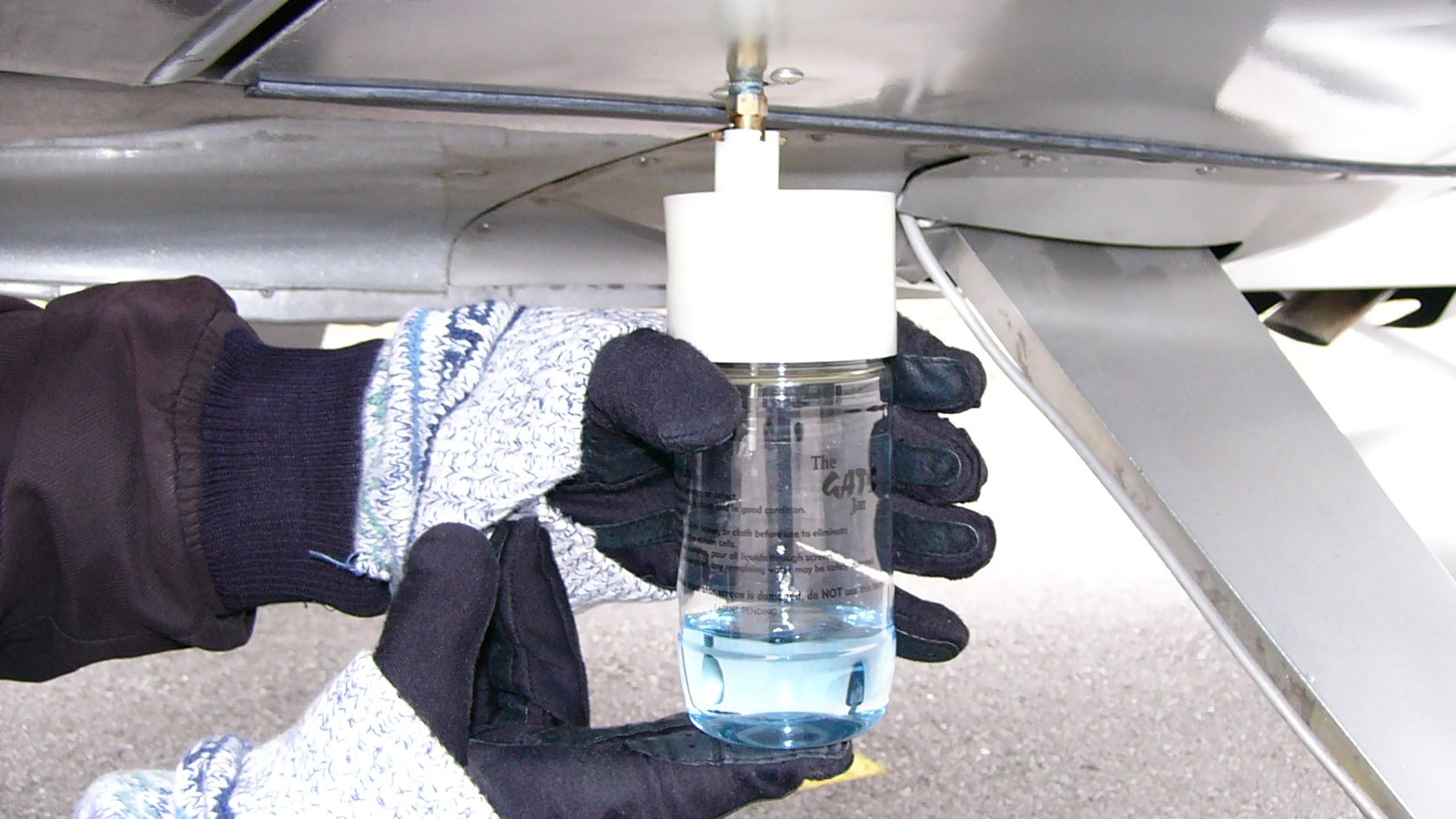 Two gloved hands holding a clear plastic sampling jar up under the wing of a light aircraft, drawing a sample from the fuel drain. The jar is part filled with pale blue liquid, the colour leaded aviation gasoline is dyed. Bare riveted metal and a strut fill the upper half of the frame and the ground is out of focus below.