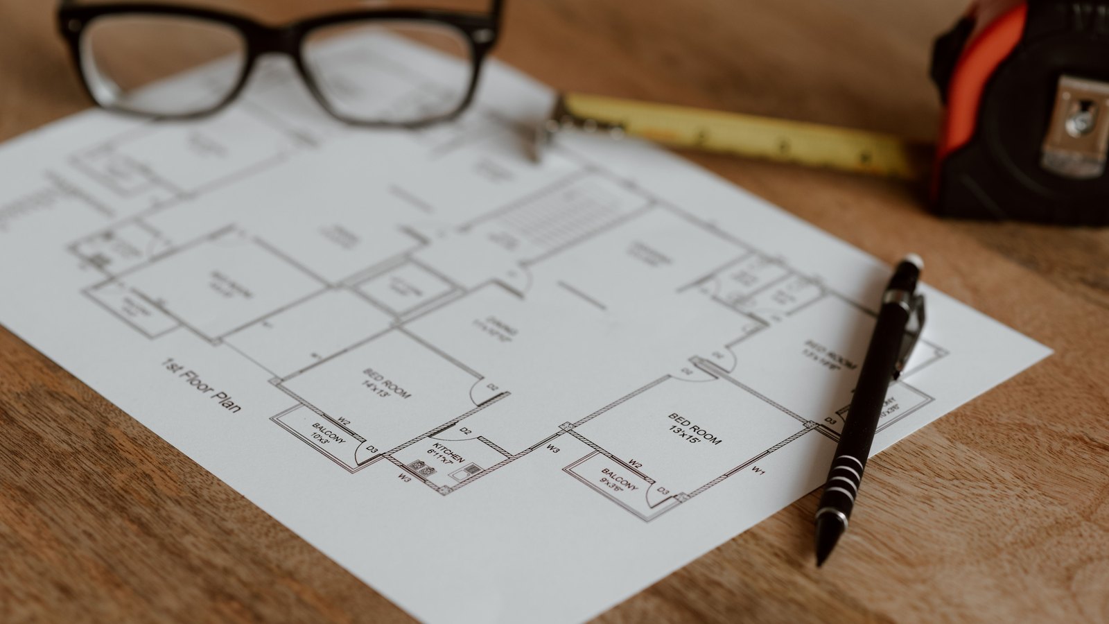 A printed architectural floor plan on a wooden table beside glasses, a pen and a tape measure, not a photograph of this event.