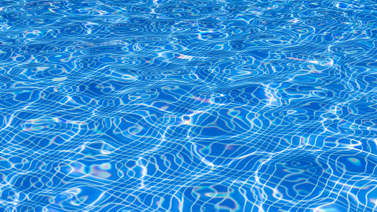 Sunlit blue water filling the frame at the surface of a swimming pool, the ripples throwing a fine white net of light across the bottom. No person, no building, no poolside, no signage and no identifying mark of any kind appears in the frame. A stock photograph, not a photograph of this event.