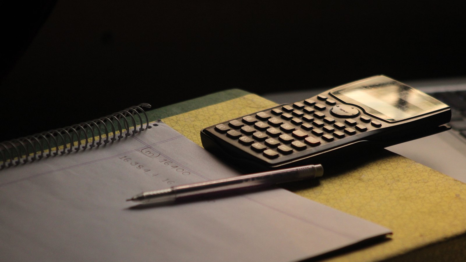A calculator, a pen and a spiral notebook of handwritten figures on a desk in low light.