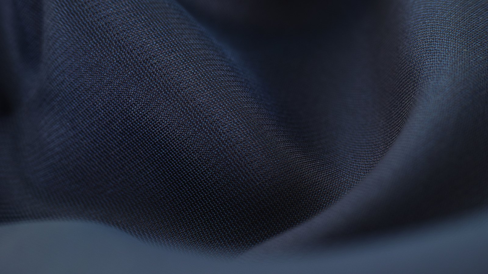 A close photograph of dark navy fabric lying in soft folds, the weave visible across a raised ridge running left to right, the light falling away into shadow at the top and a paler fold crossing the lower edge.
