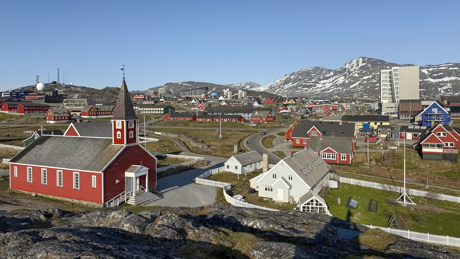 A panoramic view of Nuuk, Greenland, with colourful buildings, a red church and snow-streaked mountains behind the city.