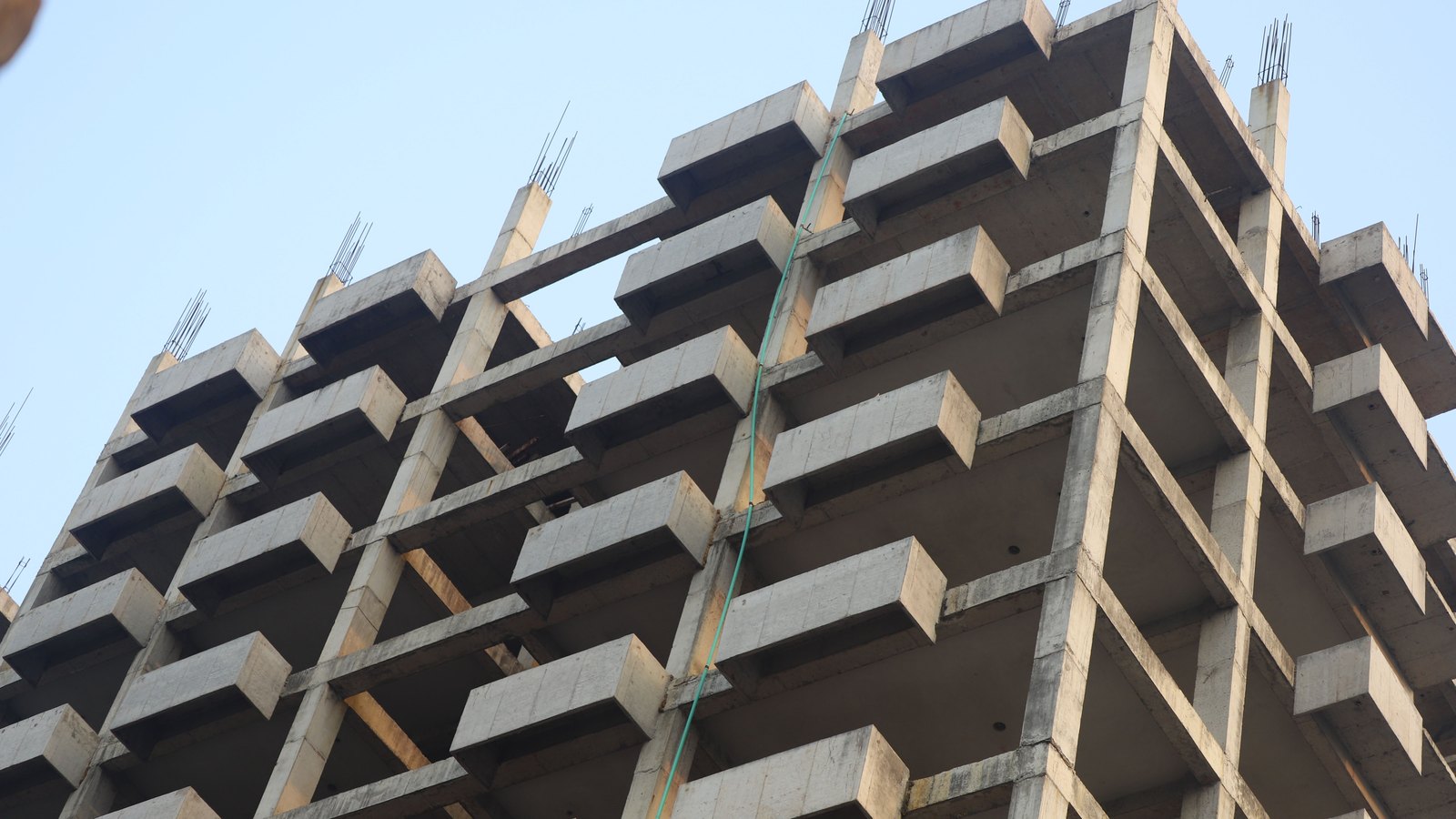 The bare concrete frame of an unfinished multi-storey building seen from below, with projecting floor slabs and balconies and lengths of reinforcing bar standing up from the tops of the columns against a pale sky. No people, machinery or signage are in the frame.