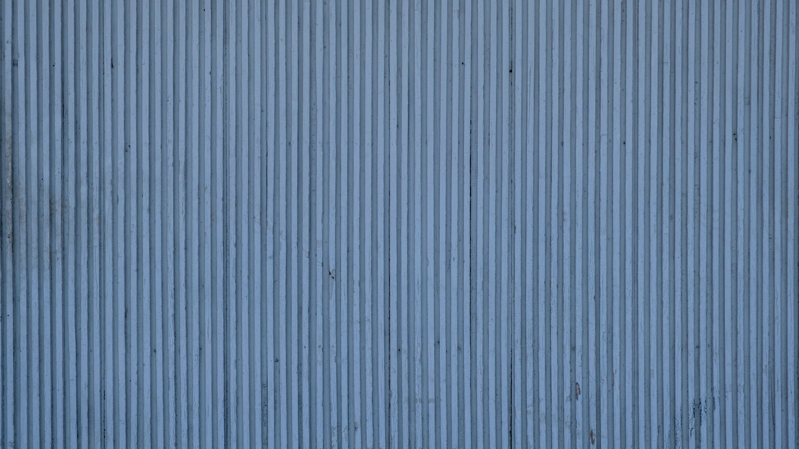 A pale blue-grey wall of fine vertical ridges running the full height of the frame, evenly spaced edge to edge, with faint darker streaks running down the surface.