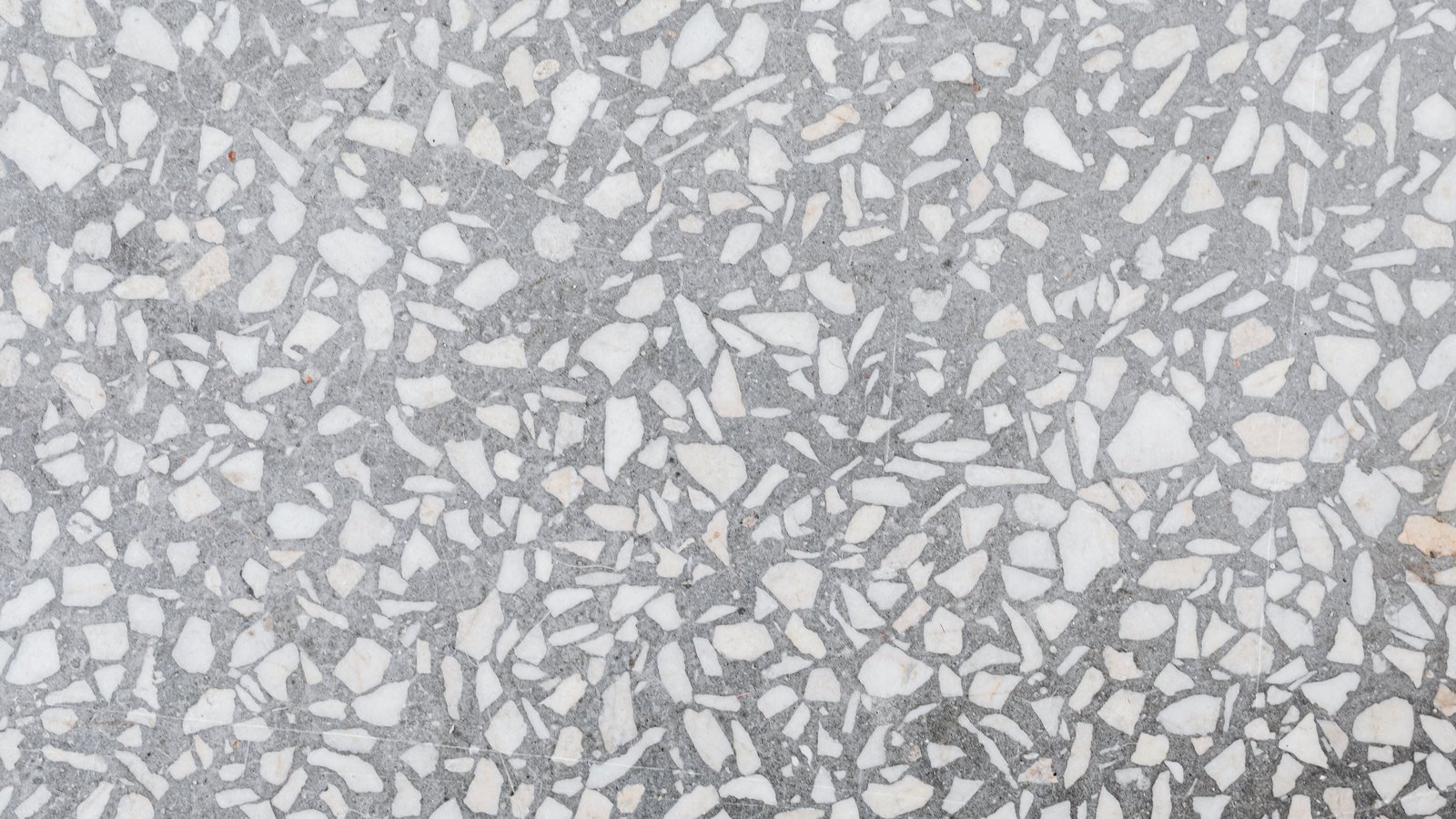 A flat slab of grey terrazzo filling the whole frame, thousands of angular white and pale cream stone chips of many different sizes set into a fine dark grey cement matrix, with a handful of faint rust-coloured flecks.