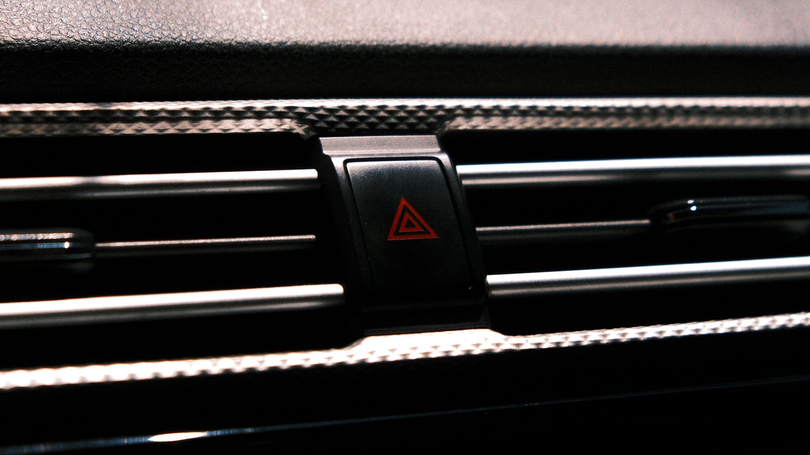 A close up of a car dashboard in low light, with the hazard warning switch at the centre of the frame carrying the standard red triangle symbol, the air vent slats and trim running across the picture on either side, and no person, number plate, place name, agency marking or legible text anywhere in the shot. A generic illustration of a driver reaching for the hazard lights on meeting blocked traffic, not a photograph of this event.