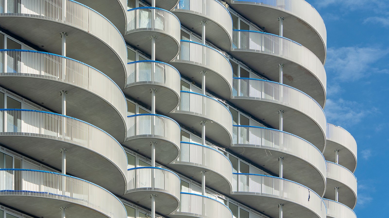 Curved concrete balconies stacked in tiers up the side of a modern residential building, each edged with a blue and white railing, photographed close against a clear blue sky. No people, signage, lettering or landmarks appear. A generic illustration of an apartment building, not a photograph of Everly or of any Naples property.