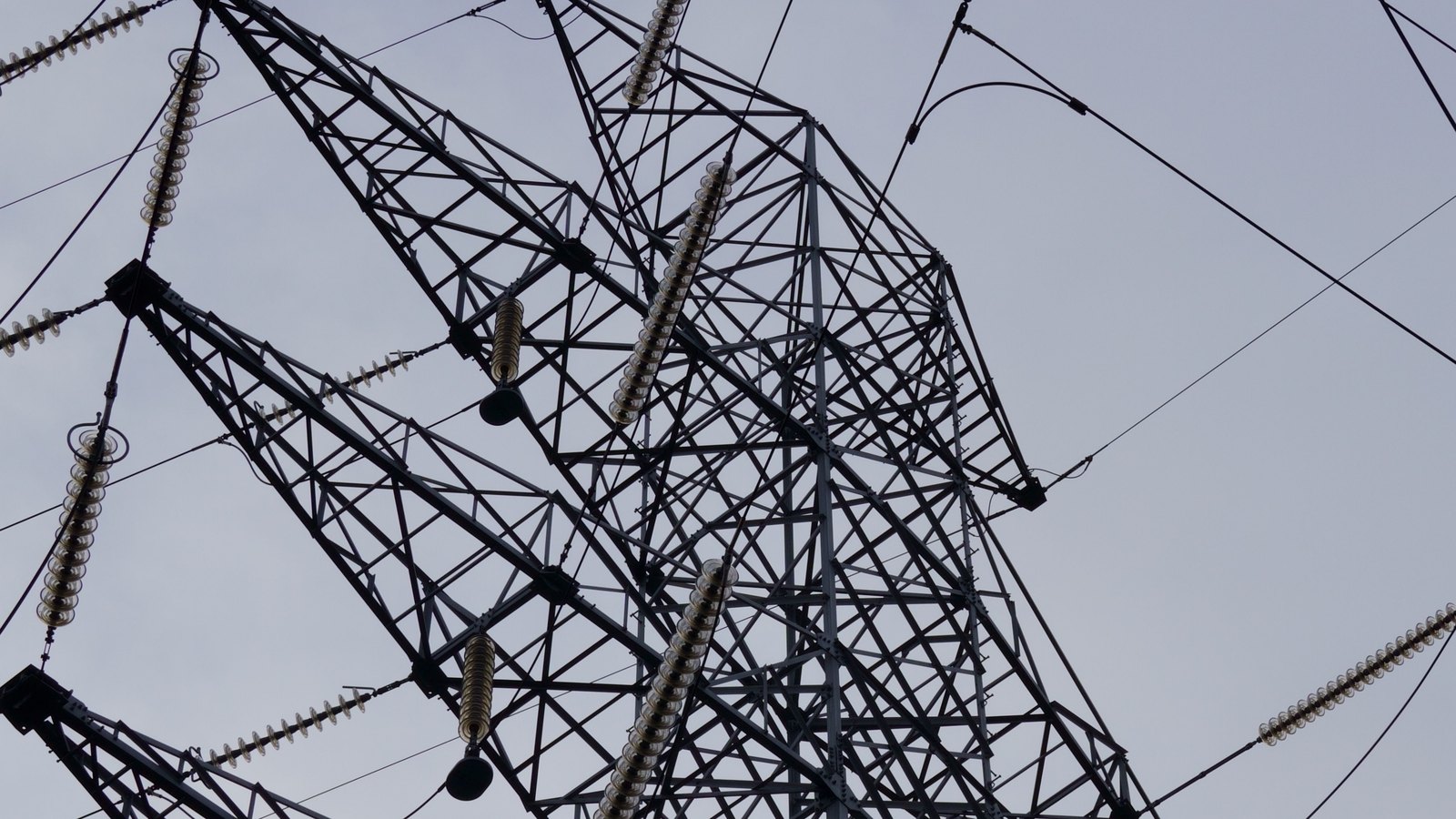A low angle photograph looking up through the arms of a steel lattice electricity transmission tower against a flat pale grey sky, with vertical and horizontal strings of glass insulator discs hanging from the crossarms and conductors running out of frame on both sides. No ground, horizon, person, marking or lettering is in view.